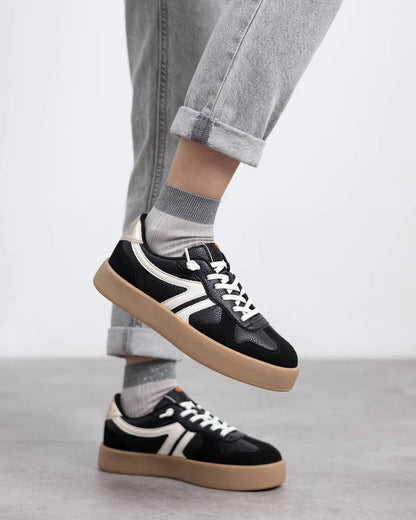 Person wearing light grey socks and fitted light grey jeans with black non-slip trainers featuring white accents and gum soles, standing on a smooth grey surface.