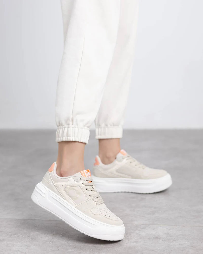 Person wearing vegan trainers with chunky soles and peach accents, paired with white joggers and featuring a removable insole, stands on a light grey floor.