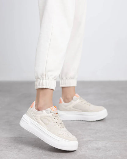 Person wearing vegan trainers with chunky soles and peach accents, paired with white joggers and featuring a removable insole, stands on a light grey floor.